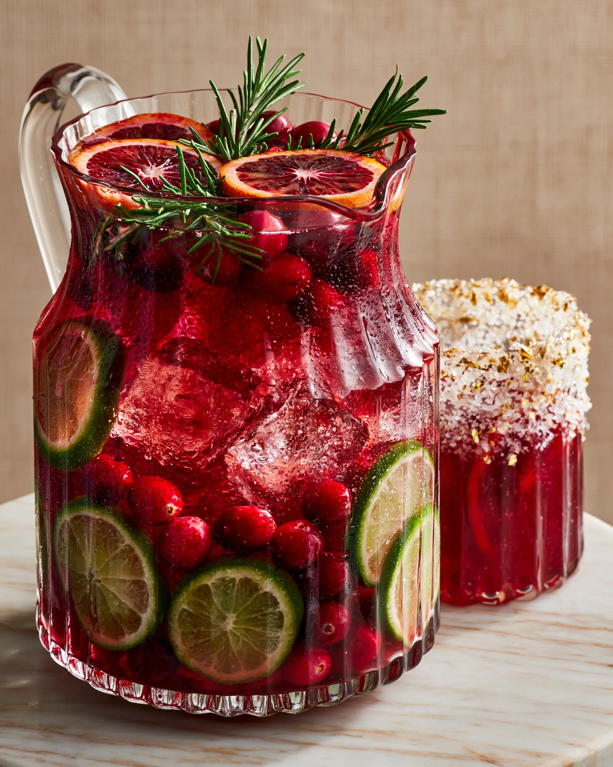 A vintage fluted glass pitcher filled with a crimson holiday margarita on a marble table with rosemary and blood orange garnishes.
